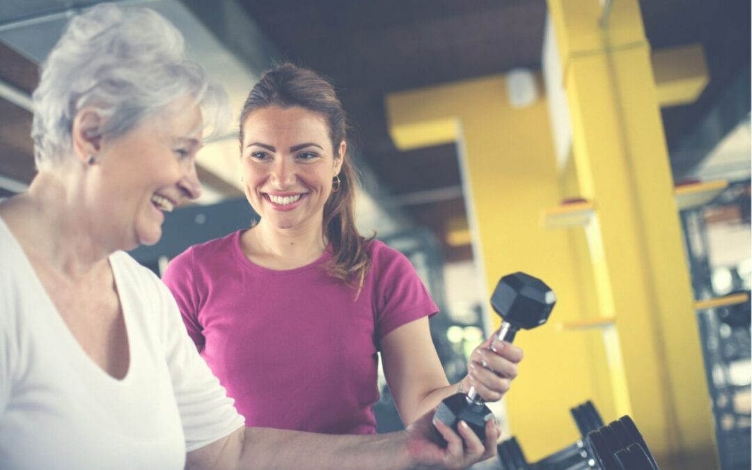 An older lady, with a bright smile on her face, stands in a gym as her coach hands her a dumbbell. She exudes enthusiasm and determination as she prepares for her strength training session. The image captures the supportive and encouraging environment of the gym, highlighting the importance of personalized coaching and guidance for individuals over 55.